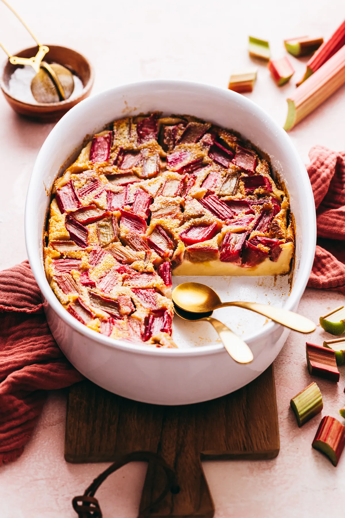 Tranche de clafouti à la rhubarbe sur assiette blanche, bords dorés et éclats de compote rosée, tige de rhubarbe en