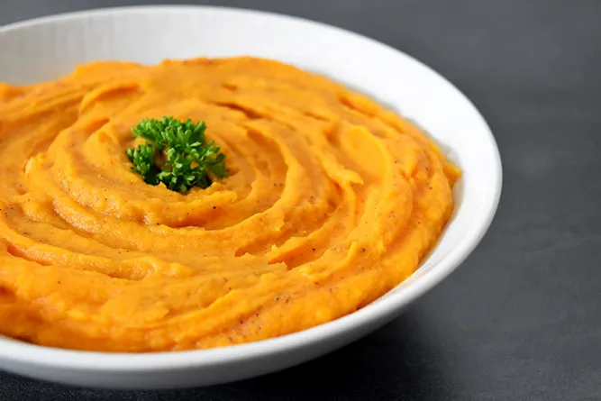 Bol en céramique contenant une purée de patate douce orange vif, nappée d'un filet d'huile d'olive et parsemée de graines et